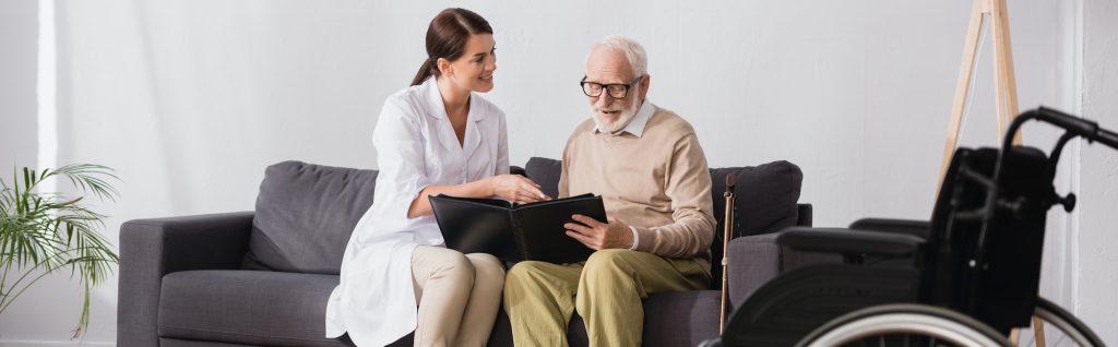 geriatric nurse and elderly man looking at photos while sitting on sofa, banner